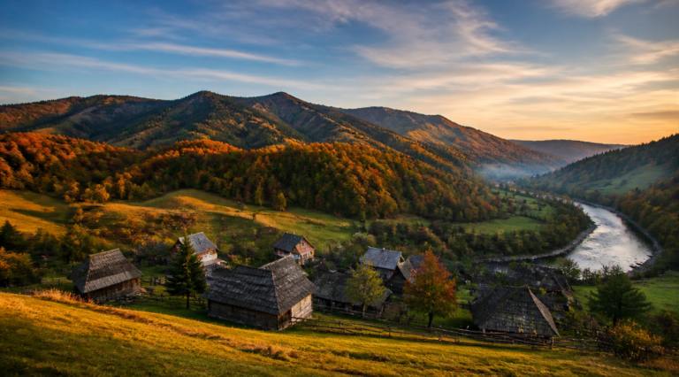 A breathtaking landscape photograph of Northern Bukovina in Ukraine, featuring the rolling Carpathian mountains covered in dense autumn forests with vibrant orange, red, and golden foliage. In the foreground, a traditional Hutsul village with wooden houses and shingled roofs sits peacefully in a valley. A winding river reflects the soft morning light. The sky is a soft gradient of pastel blue with wispy clouds. The image has cinematic lighting, warm golden hour tones, photorealistic style, high resolution, National Geographic quality, sharp details, and a wide-angle composition that captures the vastness and serenity of the region.