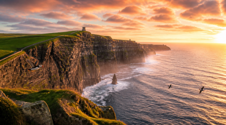 A breathtaking panoramic photograph of the Cliffs of Moher in Ireland during golden hour, with dramatic 214-meter vertical cliffs plunging into the Atlantic Ocean, lush green grass on top, ancient stone walls along the cliff edge, O'Brien's Tower visible on the headland, scattered seabirds including puffins flying, soft mist rising from the ocean, dramatic Irish sky with golden and pink clouds, waves crashing against dark rocks below, atmospheric travel photography, high resolution, professional landscape photography style, vibrant natural colors, 4K quality
