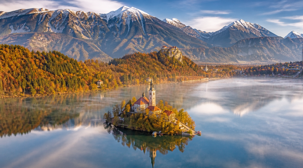 A breathtaking panoramic view of Lake Bled in Slovenia with the iconic small island featuring a baroque church and bell tower in the middle of the turquoise water, Bled Castle perched on a cliff in the background, surrounded by the majestic Julian Alps in vibrant autumn colors of orange and gold, soft morning golden light with delicate mist rising from the water surface, reflection of mountains in the calm lake, photorealistic travel photography style, ultra high detail, cinematic composition, vivid colors, 4K quality