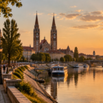 A photorealistic wide-angle view of Szeged, Hungary, at golden hour, with the iconic Votive Church in the background, the calm Tisza river in the foreground, locals and expats walking along the riverside promenade, warm tones, soft sunlight reflecting on historic architecture, cobblestone streets, gentle summer atmosphere, high detail, cinematic composition.